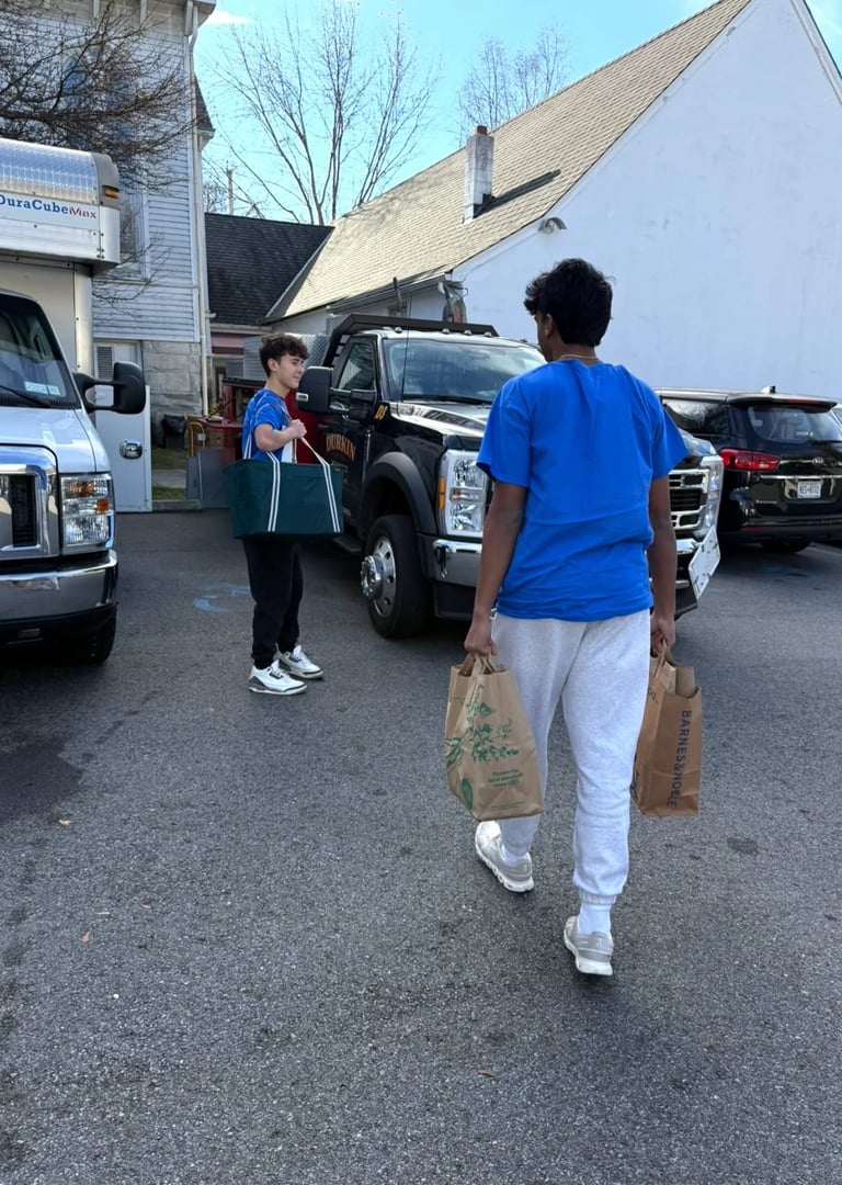 Two people in blue shirts carrying grocery bags in a parking lot near parked vehicles and storefronts