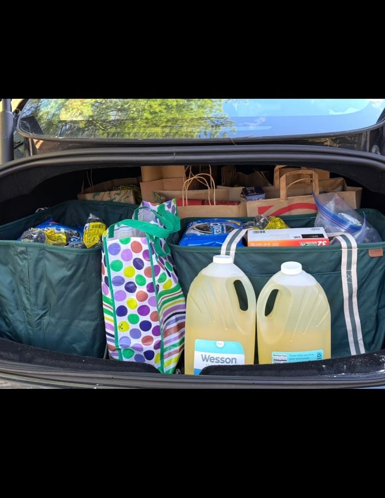 Car trunk filled with groceries, supplies, and household items including milk jugs, colorful polka dot bag, and cardboard boxes